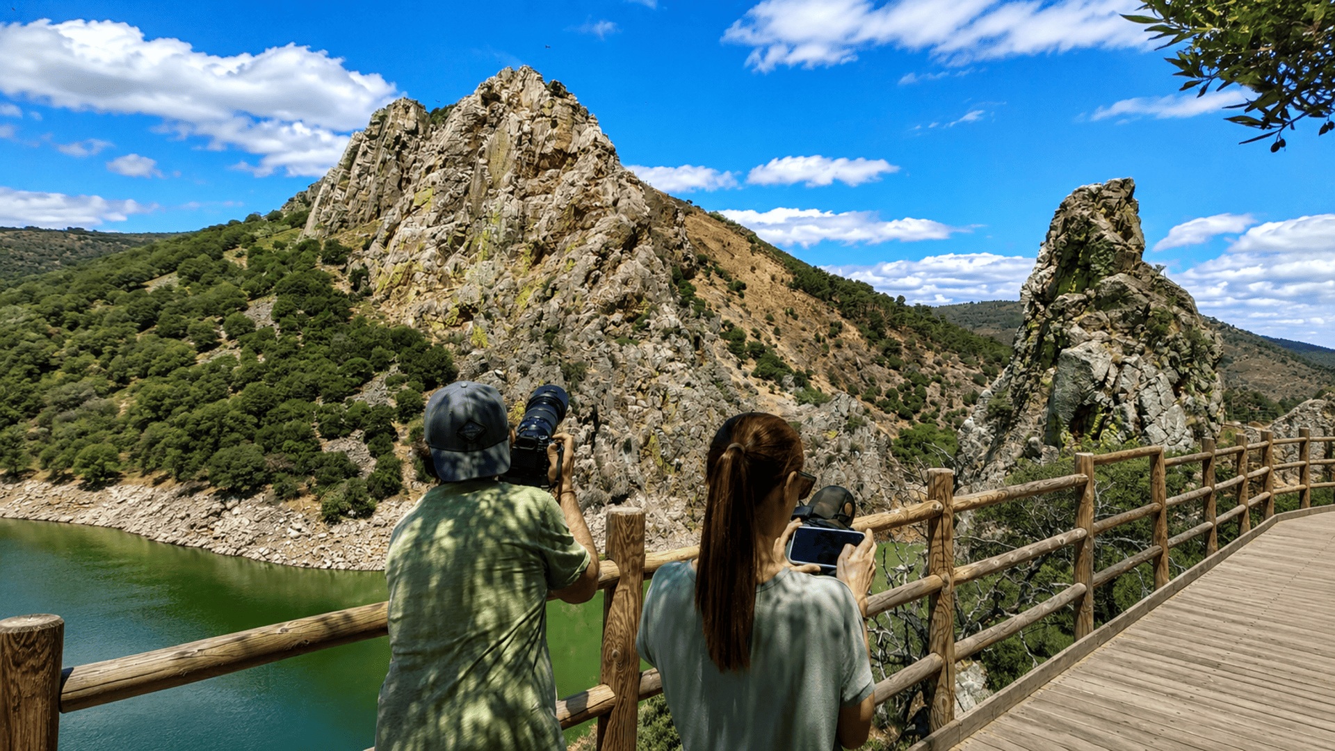 Visita_guiada_4 Dos visitantes observan y fotografían el paisaje rocoso de Monfragüe desde una pasarela de madera junto al río.